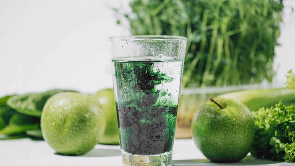 chlorophyll in a glass of water on a white background near are various fresh vegetables fruits and roots with lettuce and spinach